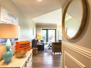 A hallway leading to a living area with a sofa, a lamp, a mirror, and a view of the outdoors.