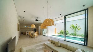 Una sala moderna con un sofá grande, una mesa de centro y un comedor con una mesa y sillas. Hay un ventanal con vistas a una piscina y zonas verdes.