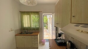 Une cuisine compacte équipée d’un évier, d’une machine à café et d’un grille-pain. Une fenêtre avec des rideaux verts offre une vue sur un jardin.