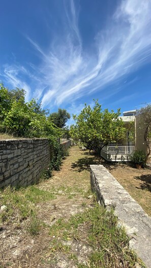 Un chemin bordé d’un mur de pierres qui mène à un bâtiment doté d’un balcon, le tout entouré de verdure et sous un ciel dégagé.