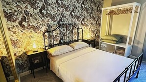 Un dormitorio con papel tapiz estampado, una cama con ropa de cama blanca, una mesita de noche con lámpara y un armario con espejo.