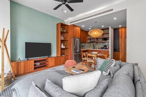 A modern living room with a large flat-screen TV, wooden cabinets, a dining area with a table and chairs, and a kitchen with a microwave and refrigerator.