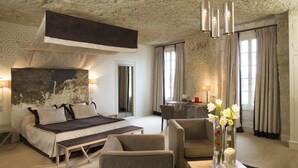 A bedroom with a large bed, bedside tables, a desk, and a chair. There is a window with curtains, a stone ceiling, and a vase with flowers on the desk. - Les Hautes Roches (Rochecorbon)