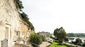 A stone building with a staircase leading up to a door, a pathway, and a view of a river and greenery. - Les Hautes Roches (Rochecorbon)