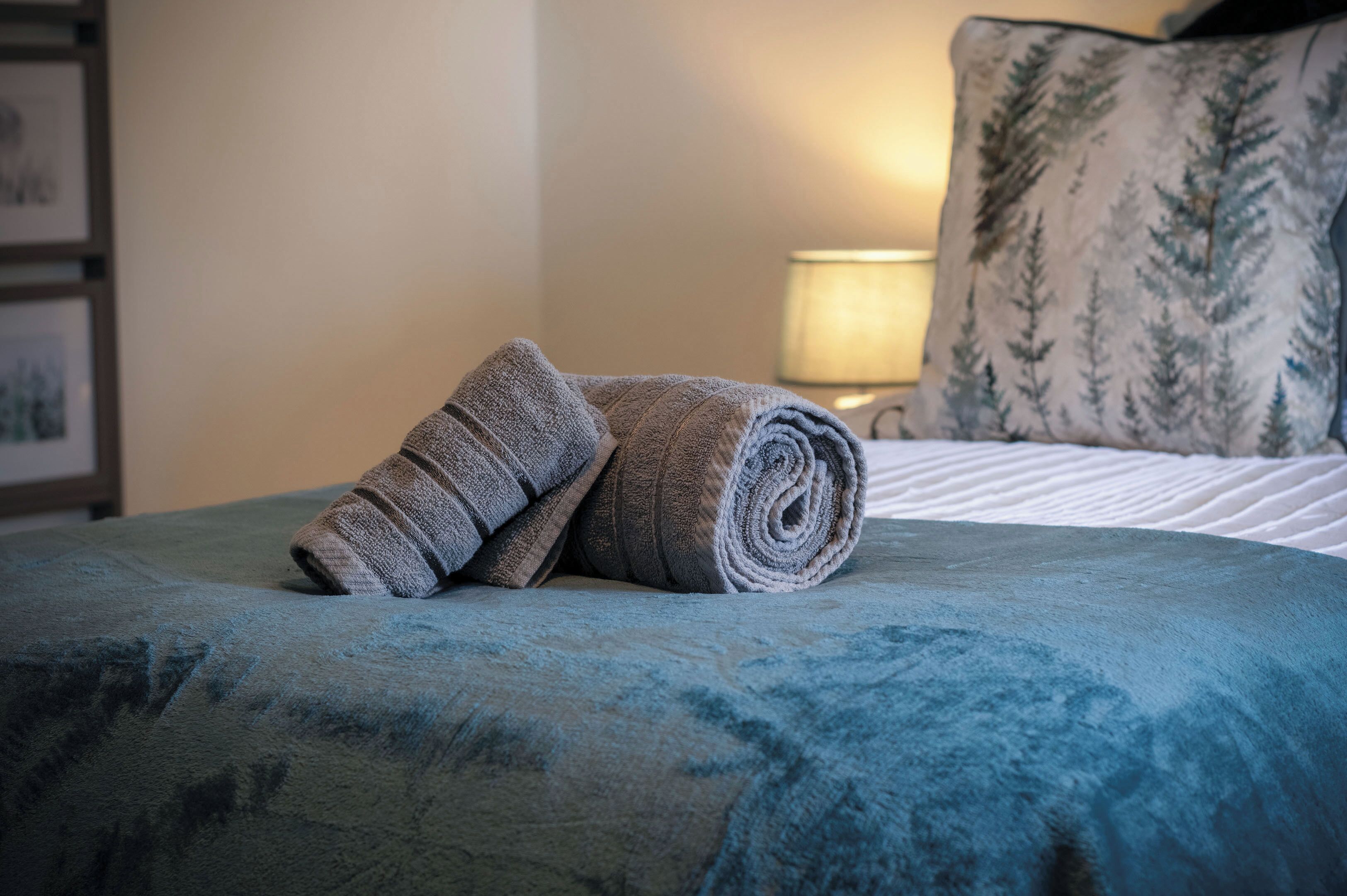 A neatly made bed with a blue bedspread and a rolled-up towel on it.