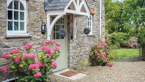 A stone house with a green door, a white window, and a small porch with hanging flower baskets. - The Stables - 2 Bedroom Cottage - Princes Gate (Narberth)