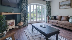 A living room with a TV, a sofa, a coffee table, and a large window with curtains. - The Stables - 2 Bedroom Cottage - Princes Gate (Narberth)