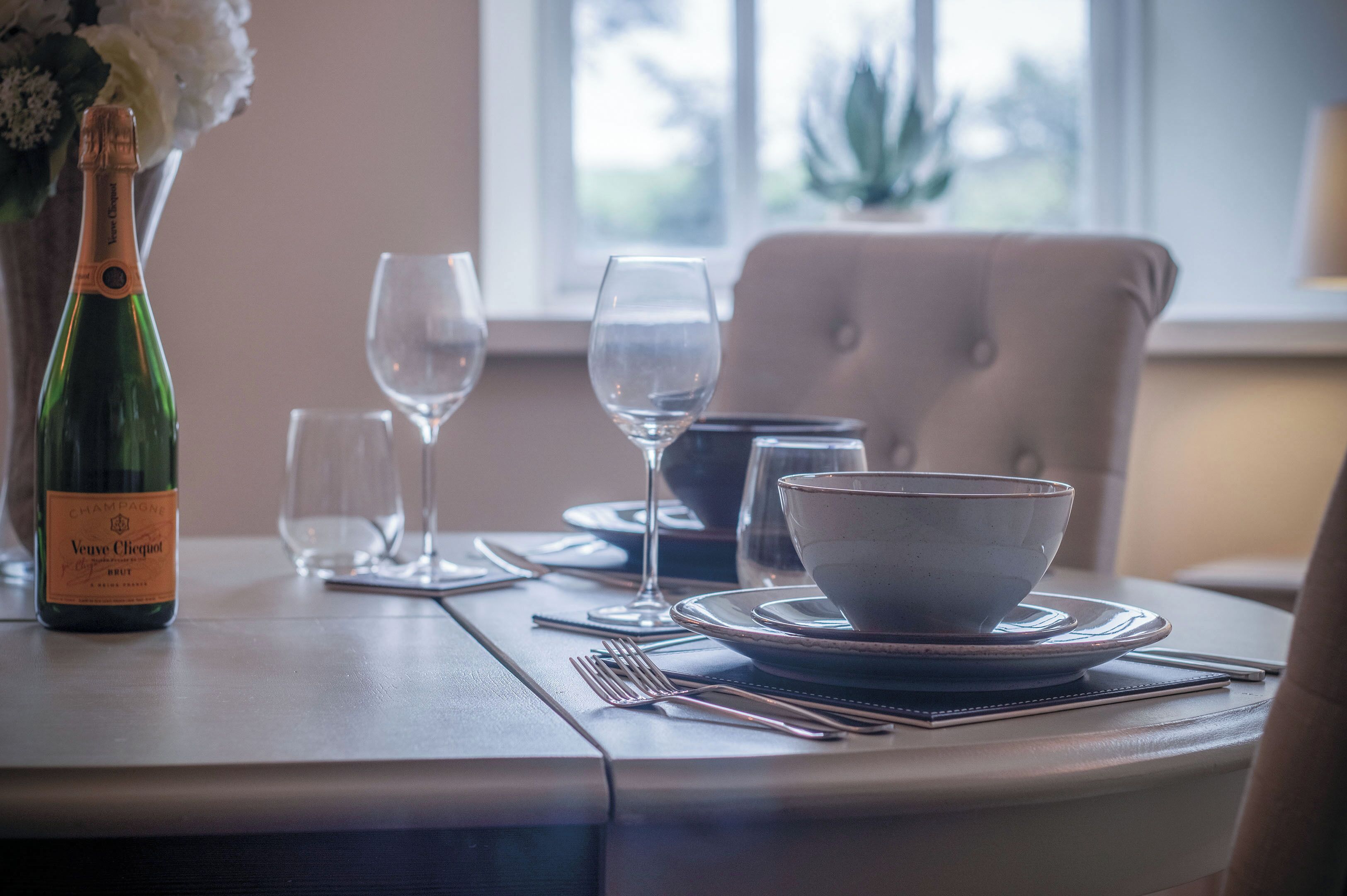 Una mesa de comedor dispuesta con una botella de Veuve Clicquot Brut champagne, dos copas de vino, un cuenco y cubiertos.