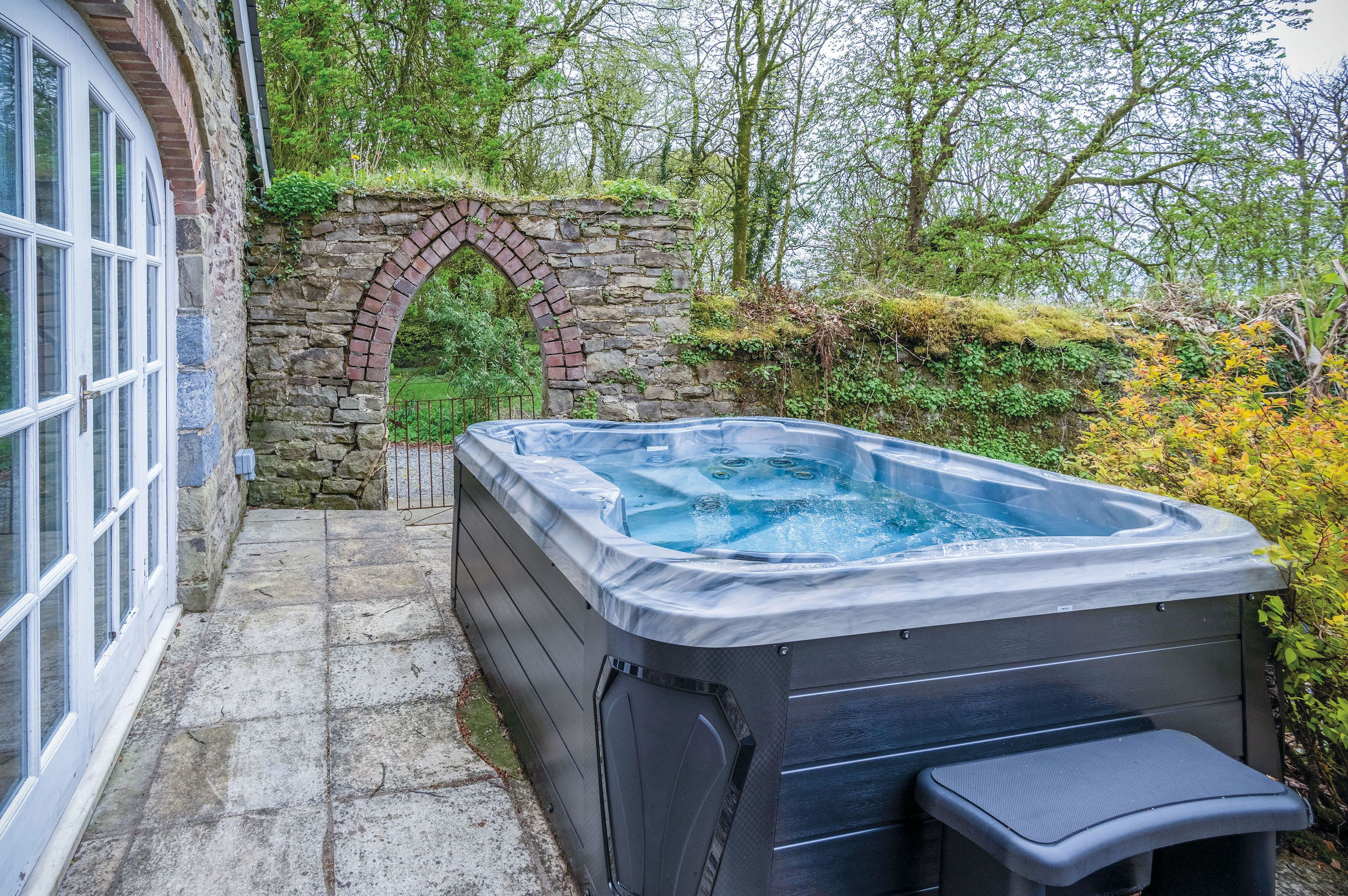 A hot tub in a garden with a stone archway and a stone wall.