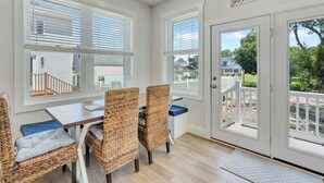 A dining area with wicker chairs and a table, large windows with blinds, and a view of a deck and house outside. - High Tide Hideaway by Seaside Vacations & Sales (Chincoteague Island)