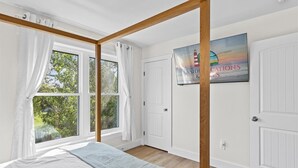 A bedroom with a four-poster bed, large windows with curtains, a door, and a wall-mounted sign. - High Tide Hideaway by Seaside Vacations & Sales (Chincoteague Island)