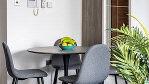 Un espace repas moderne avec une table ronde, des chaises recouvertes d’un tissu gris, un saladier rempli de fruits et une plante en pot.