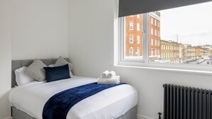 Un lit bien fait, avec une couverture bleue et du linge de lit blanc, une fenêtre donnant sur des immeubles et un radiateur au mur.