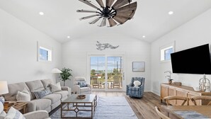 Un salon spacieux doté d’un grand ventilateur de plafond, un coin repas avec des chaises en bois, un téléviseur fixé au mur et un balcon avec vue.
