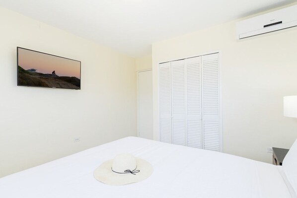 Une chambre d’hôtel avec un lit, un téléviseur fixé au mur, une armoire blanche et un chapeau blanc posé sur le lit.