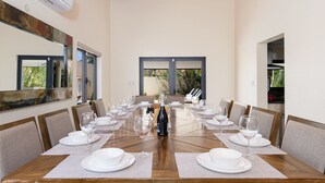 A dining room with a long wooden table set for a meal, large windows, and a modern kitchen visible through an open doorway.