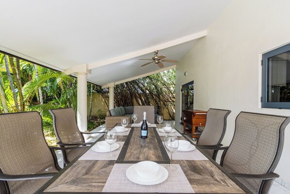 A covered outdoor dining area with a table set for six, surrounded by chairs, and a ceiling fan above.