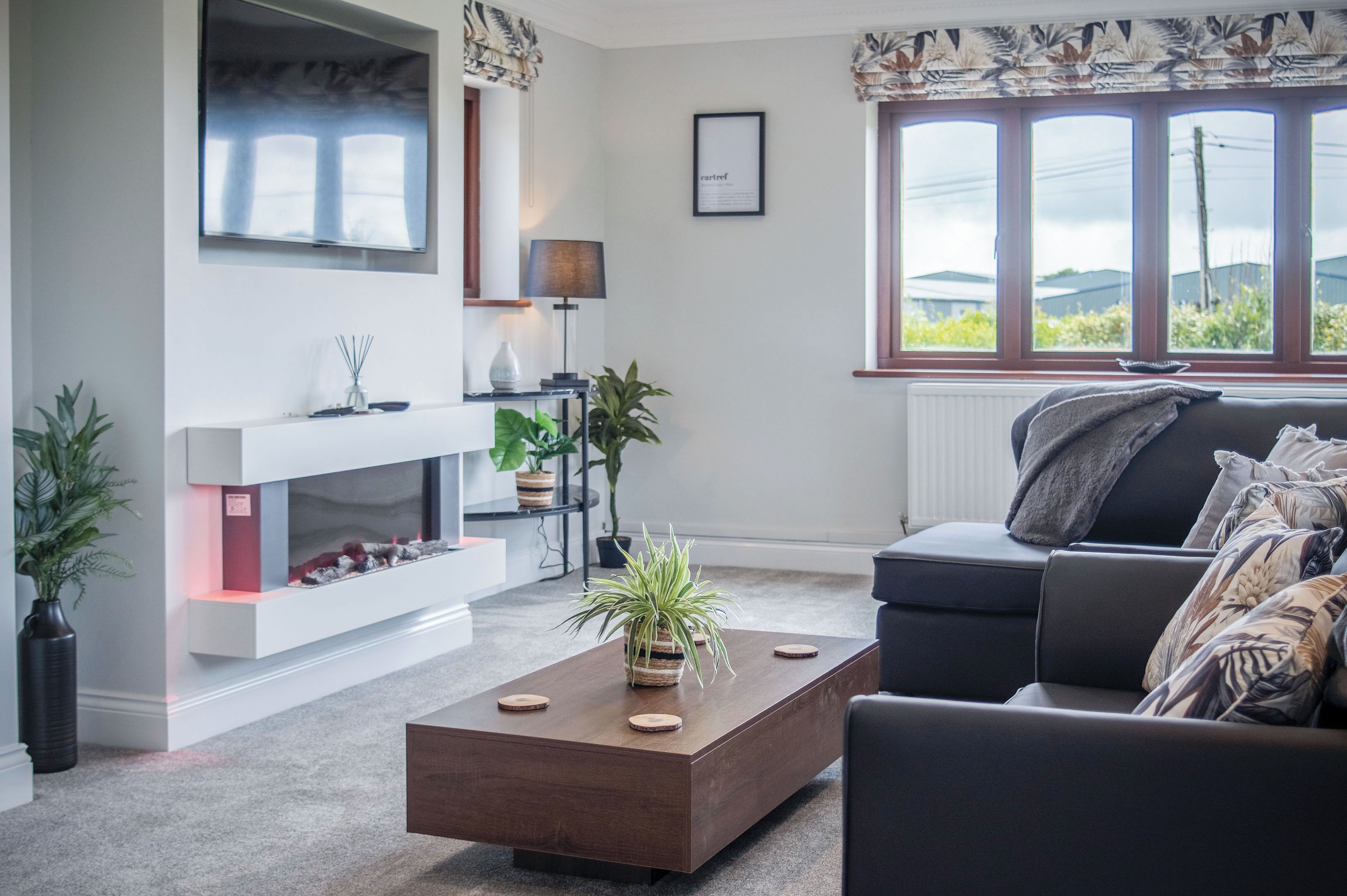 Ein modernes Wohnzimmer mit Kamin, einer Couch, einem Couchtisch und großen Fenstern mit Blick auf Grünflächen.