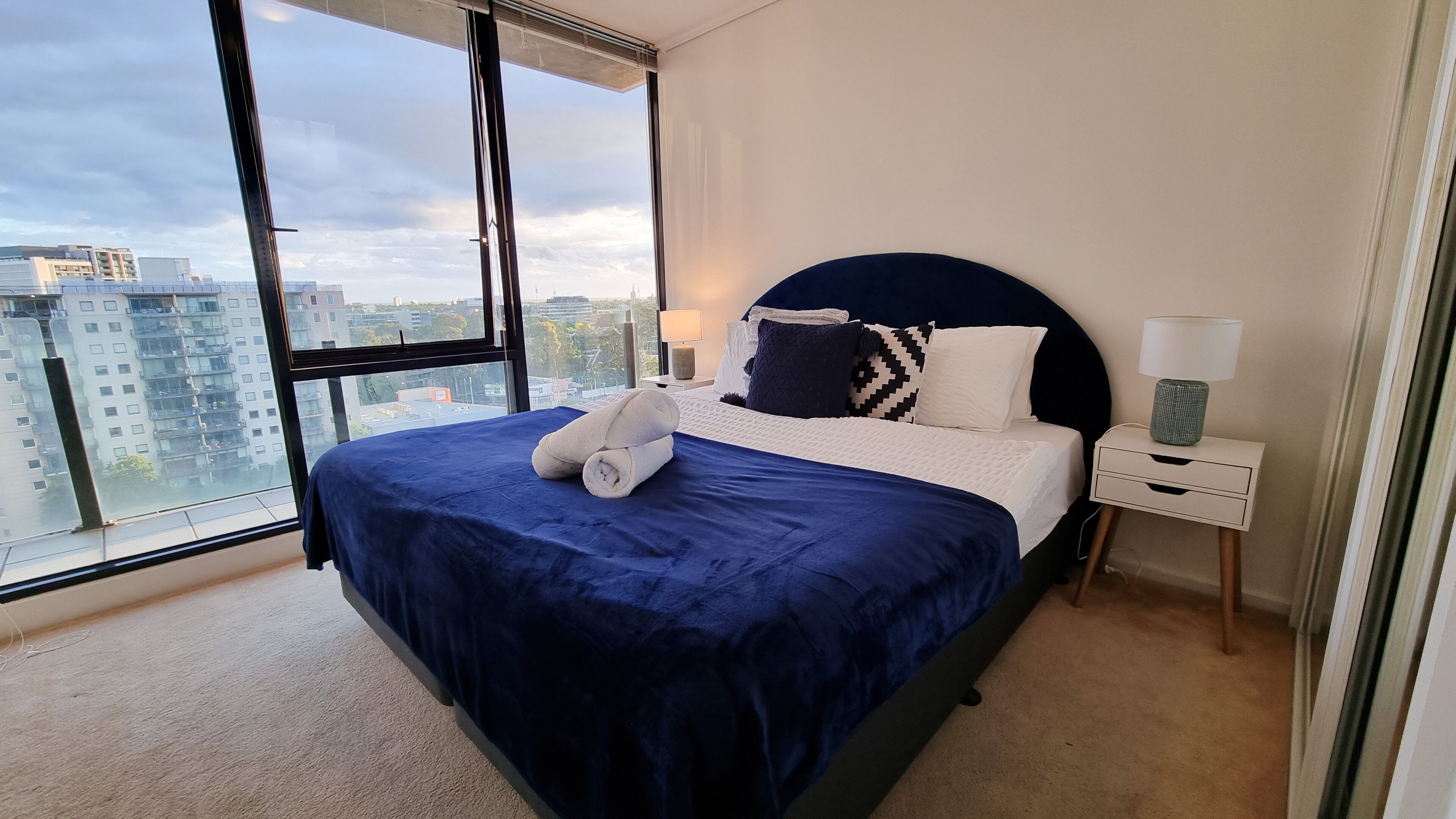 A modern bedroom with a large bed, a nightstand, a lamp, and a view of the city through large windows.