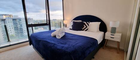 A modern bedroom with a large bed, a nightstand, a lamp, and a view of the city through large windows.