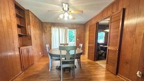 Une pièce aux murs en bois, avec un ventilateur de plafond, une table à manger entourée de chaises et une porte donnant sur une autre pièce.