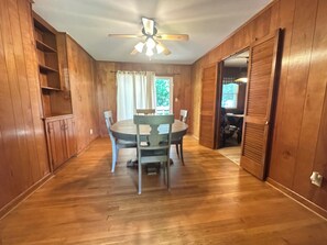 A room with wooden walls, a ceiling fan, a dining table with chairs, and a door leading to another room.