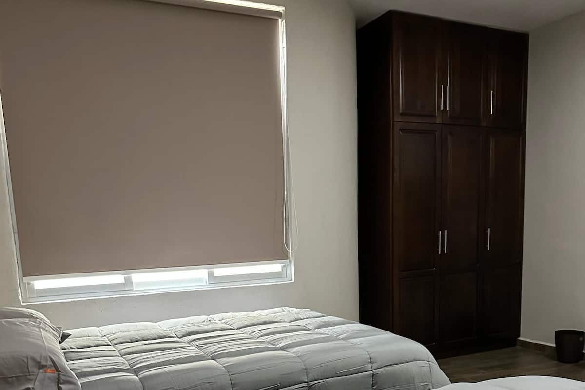 A bedroom with two beds, a window with blinds, and a dark wooden wardrobe.