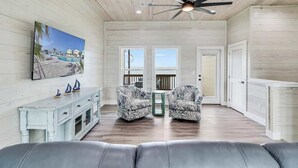 Un vaste espace de vie agrémenté d’un ventilateur de plafond, d’un tableau représentant une scène de plage, de deux fauteuils à motifs, d’une petite table d’appoint et d’un plancher en bois.