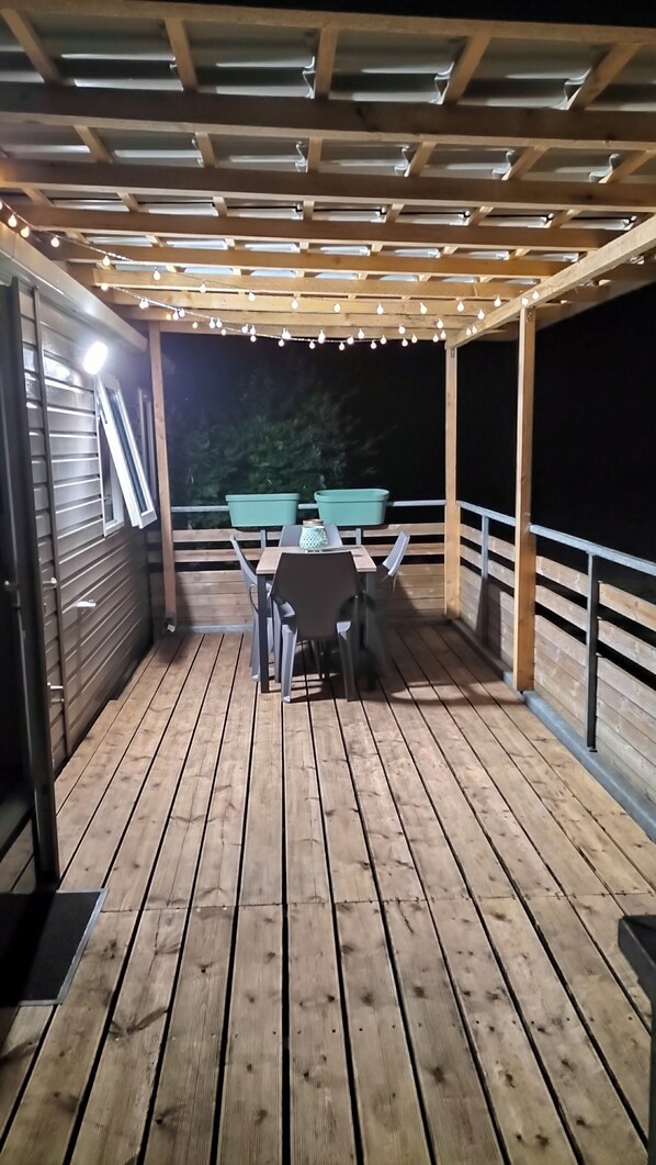 Een houten terras buiten met een tafel en stoelen, slingers erboven en uitzicht op de nachthemel.