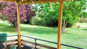 A wooden deck with a table and chairs, a pergola with string lights, and a view of a grassy yard with trees. - Cottage Cosy Aux Lauriers à Lacroix sur Meuse (Lacroix-sur-Meuse)