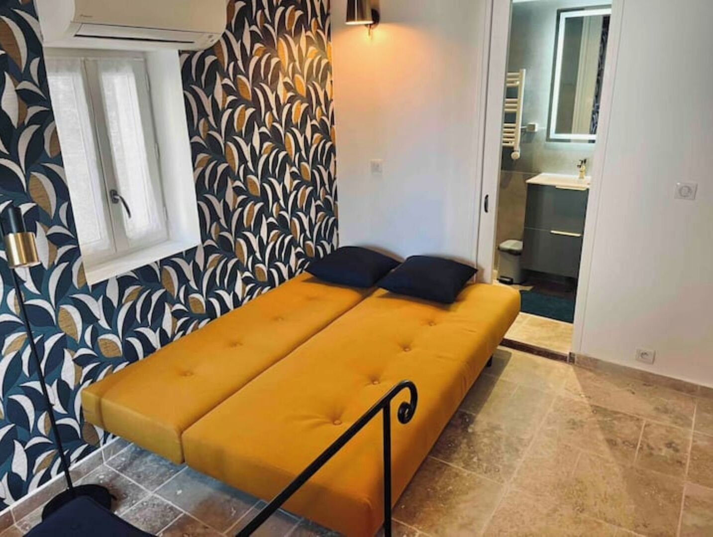Une petite chambre avec un lit simple, un mur décoré de motifs, une fenêtre et une salle de bain visible à travers une porte ouverte.