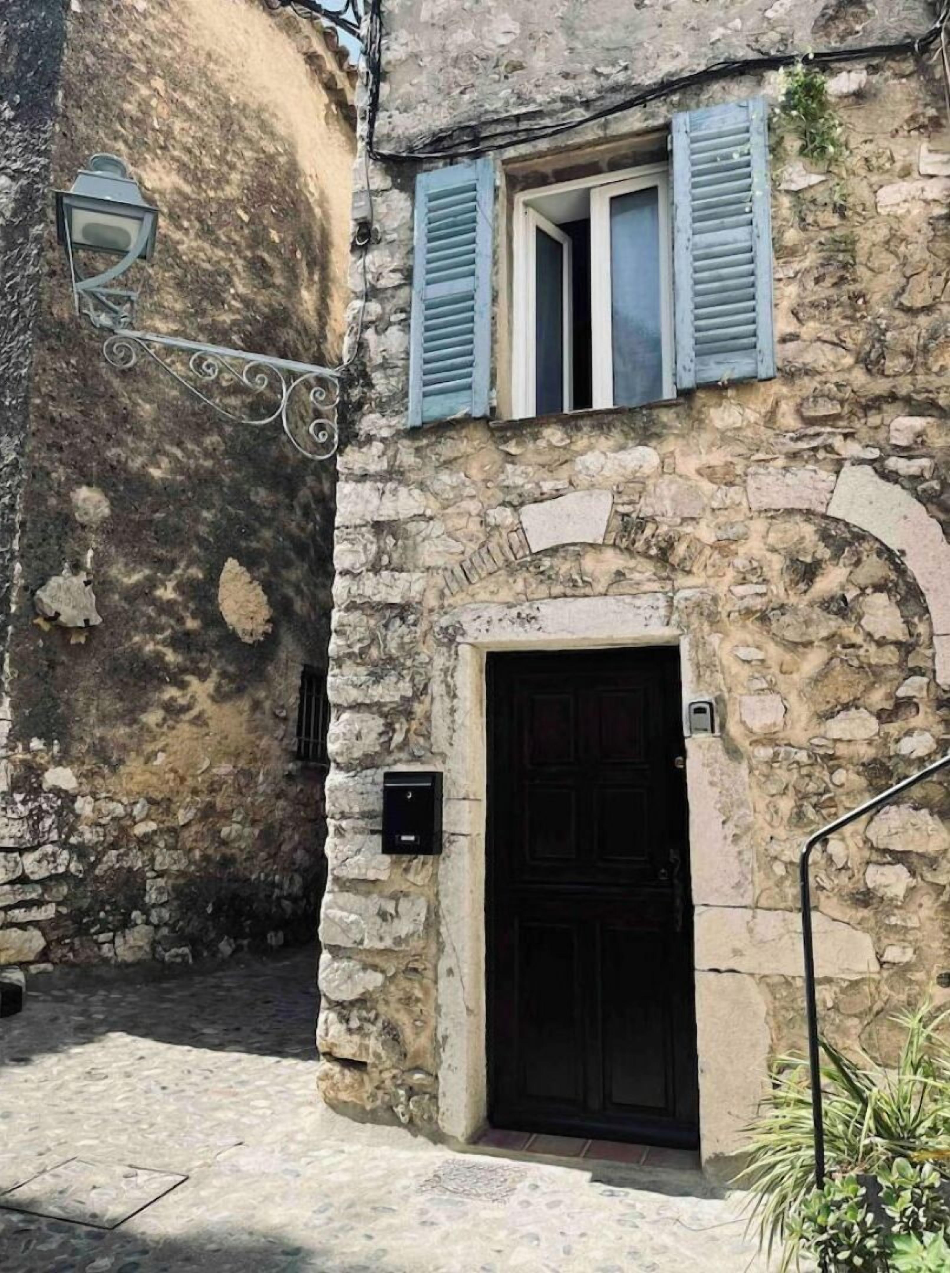 A stone building with a dark wooden door, blue shutters, and a metal lantern.