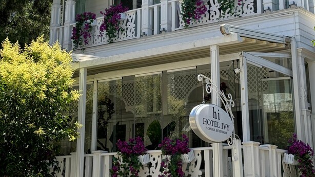 Un bâtiment à deux étages, doté de balcons ornés de fleurs violettes, d’une enseigne indiquant qu’il s’agit de l’hôtel Ivy, et d’une entrée en verre.