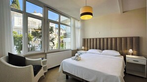 Une chambre d’hôtel avec une grande fenêtre, un lit avec du linge de maison blanc, une table de chevet avec une lampe et une chaise avec un coussin.