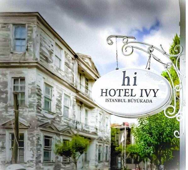 A sign for 'hi HOTEL IVY ISTANBUL BÜYÜKADA' with a classical building in the background. - hi Hotel Ivy Buyukada (Adalar)