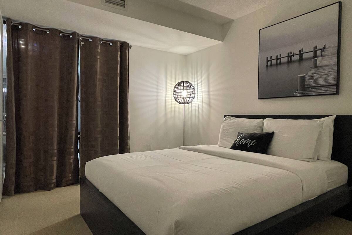 A bedroom with a large bed, a floor lamp, a painting on the wall, and a window with curtains.