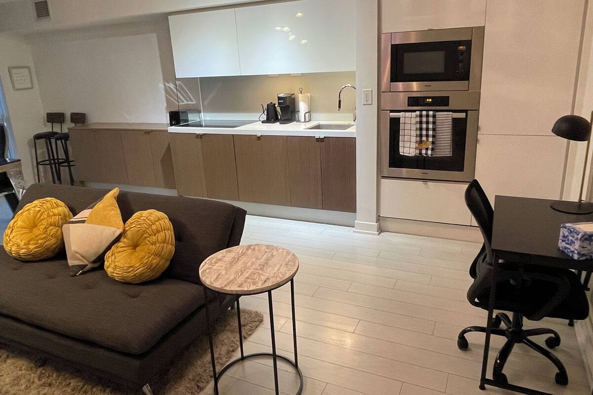 A modern living room with a sofa, a small round table, a black office chair, and a kitchen area with a microwave and oven.