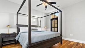 A four-poster bed with a dark frame, a light blue bedspread, and a white pillow. There is a nightstand with a lamp and a drawer. The room has wooden flooring and a ceiling fan.