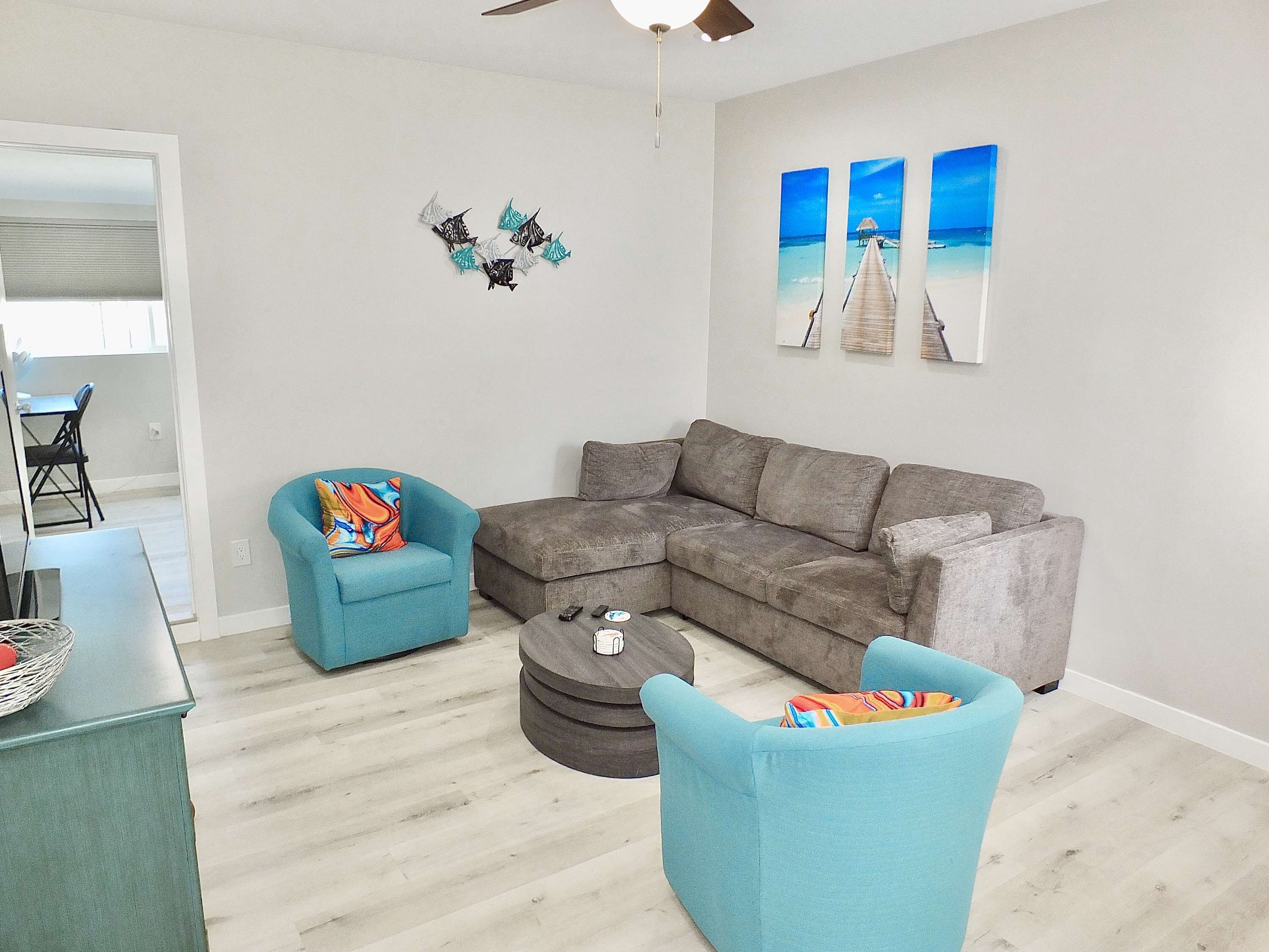 A living room with a grey sectional sofa, a blue armchair, and a round coffee table. There are three wall-mounted paintings depicting a beach scene.