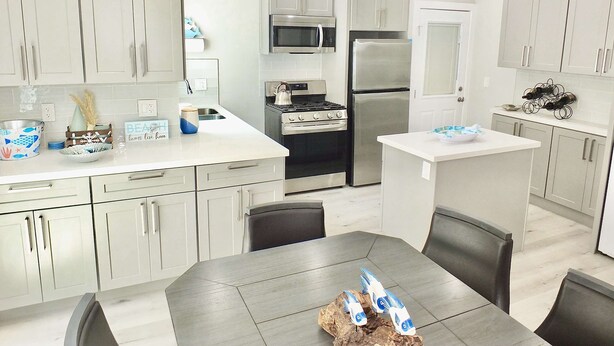 A modern kitchen with white cabinets, stainless steel appliances, and a dining area with a table and chairs. - New Beach & Bay Retreat Full of Amenities! (Long Beach)