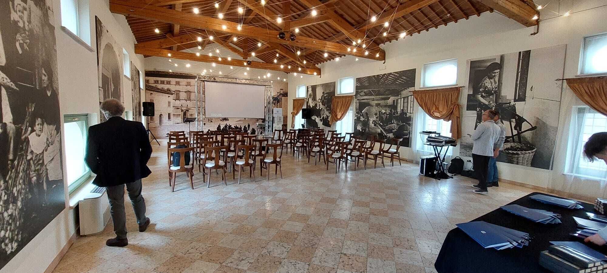 Un salón amplio con techo alto y vigas de madera, que cuenta con un escenario con instrumentos musicales y sillas dispuestas para el público.