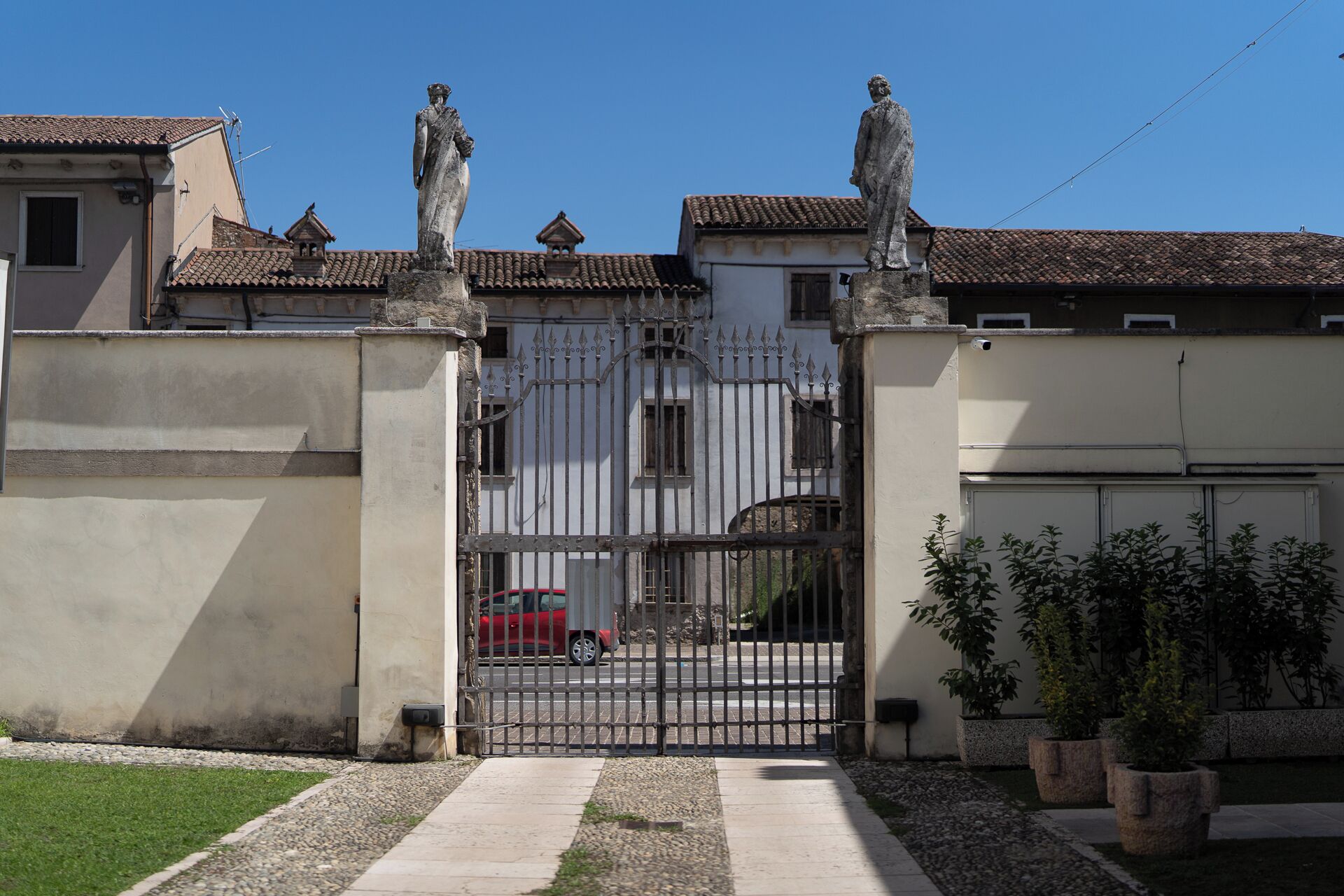 A gated entrance with two statues on top, a metal gate, and a pathway leading to a building with a tiled roof.
