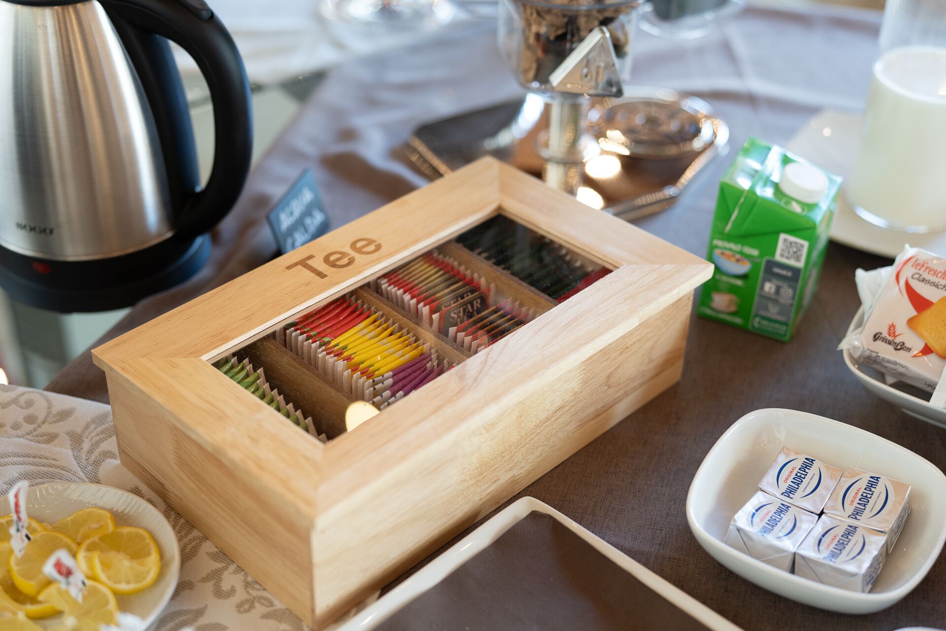 A wooden box labeled "Tee" containing a variety of tea packets, a kettle, a carton of milk, and a bowl of lemon slices on a table.