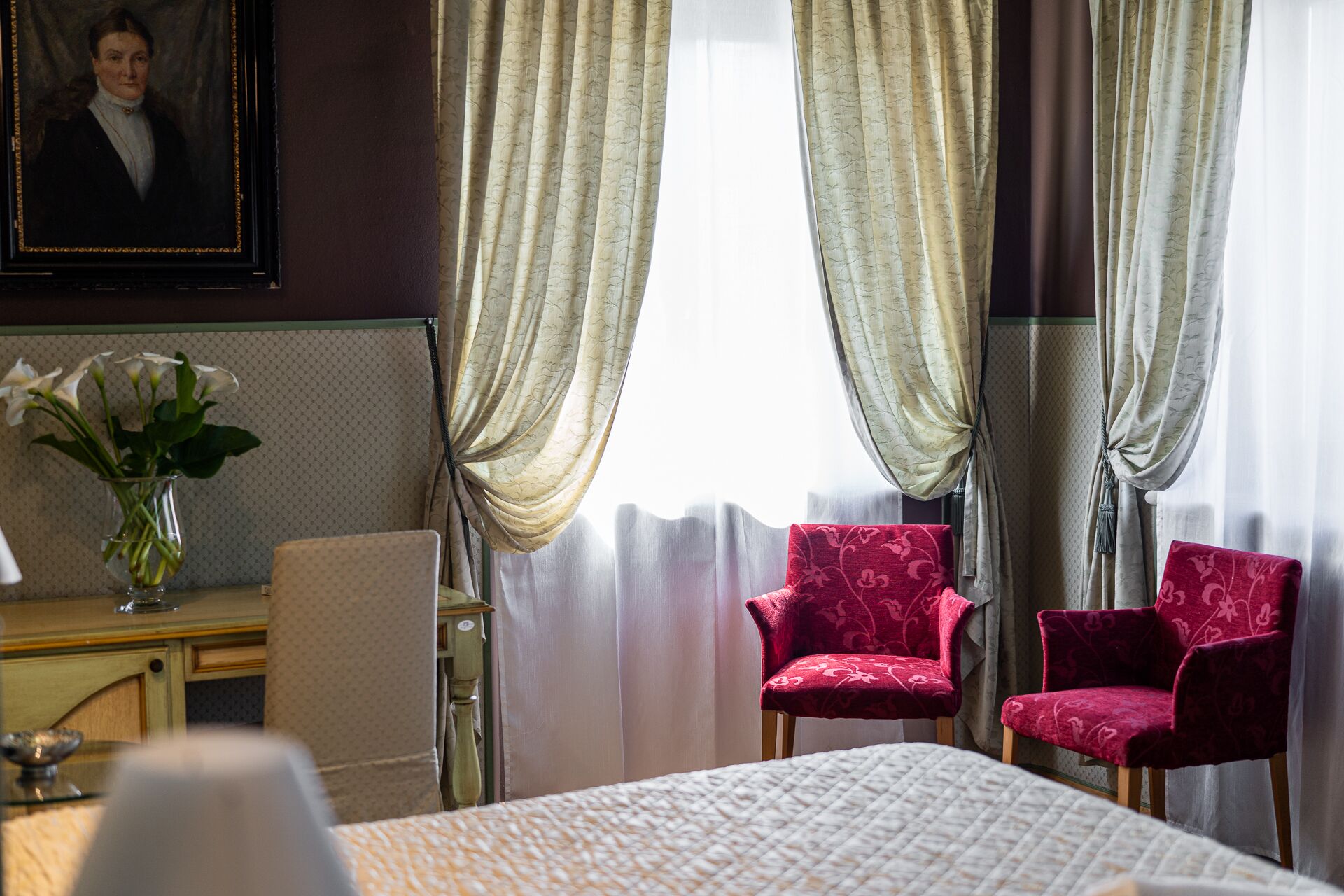 Quarto de hotel com uma cama, duas poltronas vermelhas, um vaso com flores brancas e um quadro na parede.