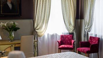 Quarto de hotel com uma cama, duas poltronas vermelhas, um vaso com flores brancas e um quadro na parede.
