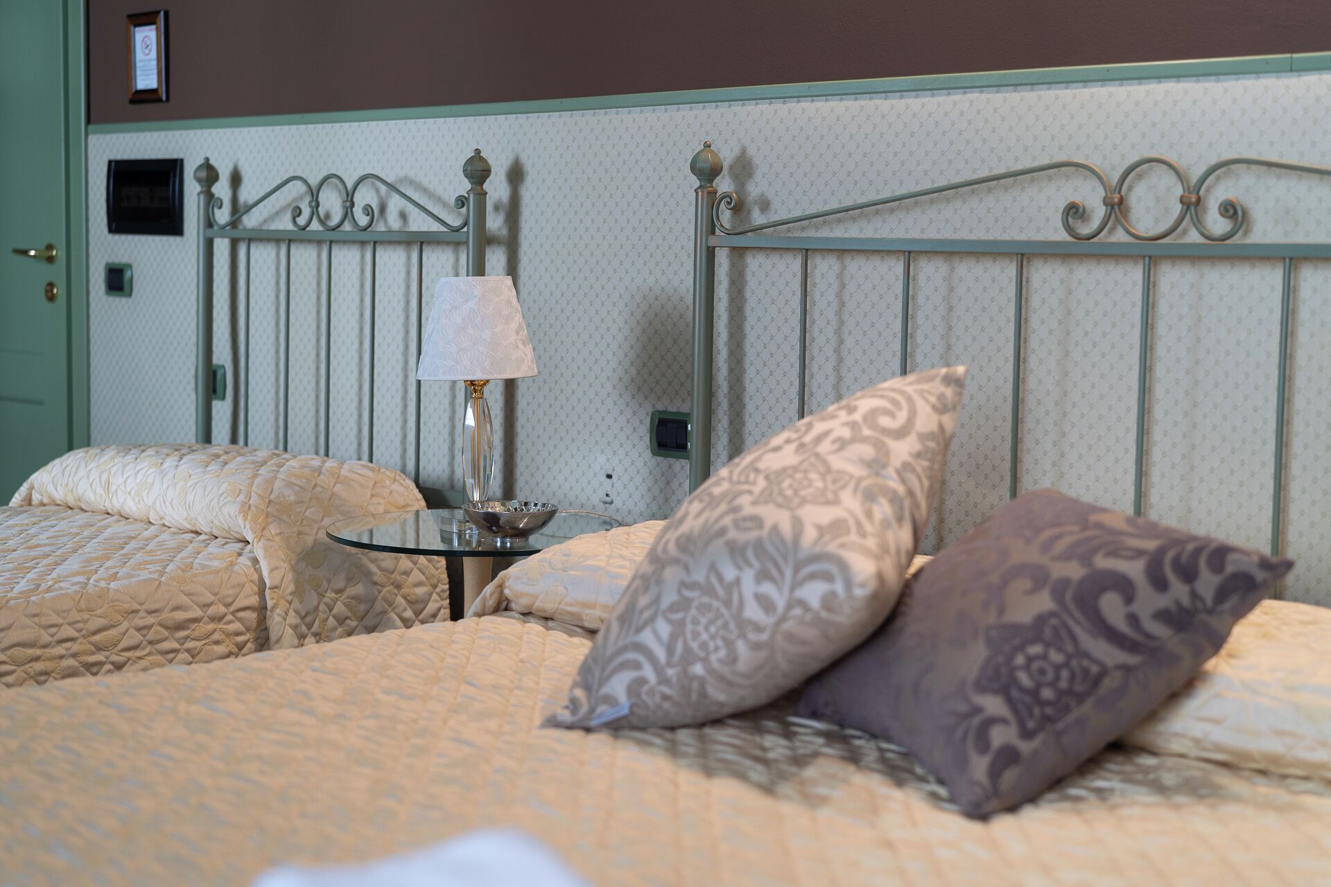 A neatly made bed with patterned pillows, a bedside table with a lamp, and a wall-mounted light switch.