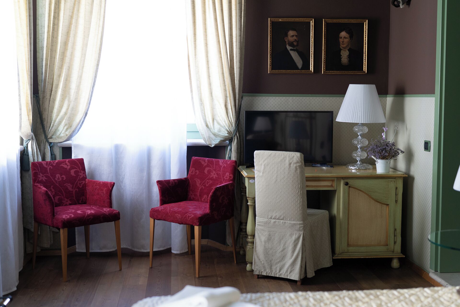 Una habitación con dos sillones rojos, un escritorio de madera con una lámpara, un televisor y dos retratos enmarcados en la pared.