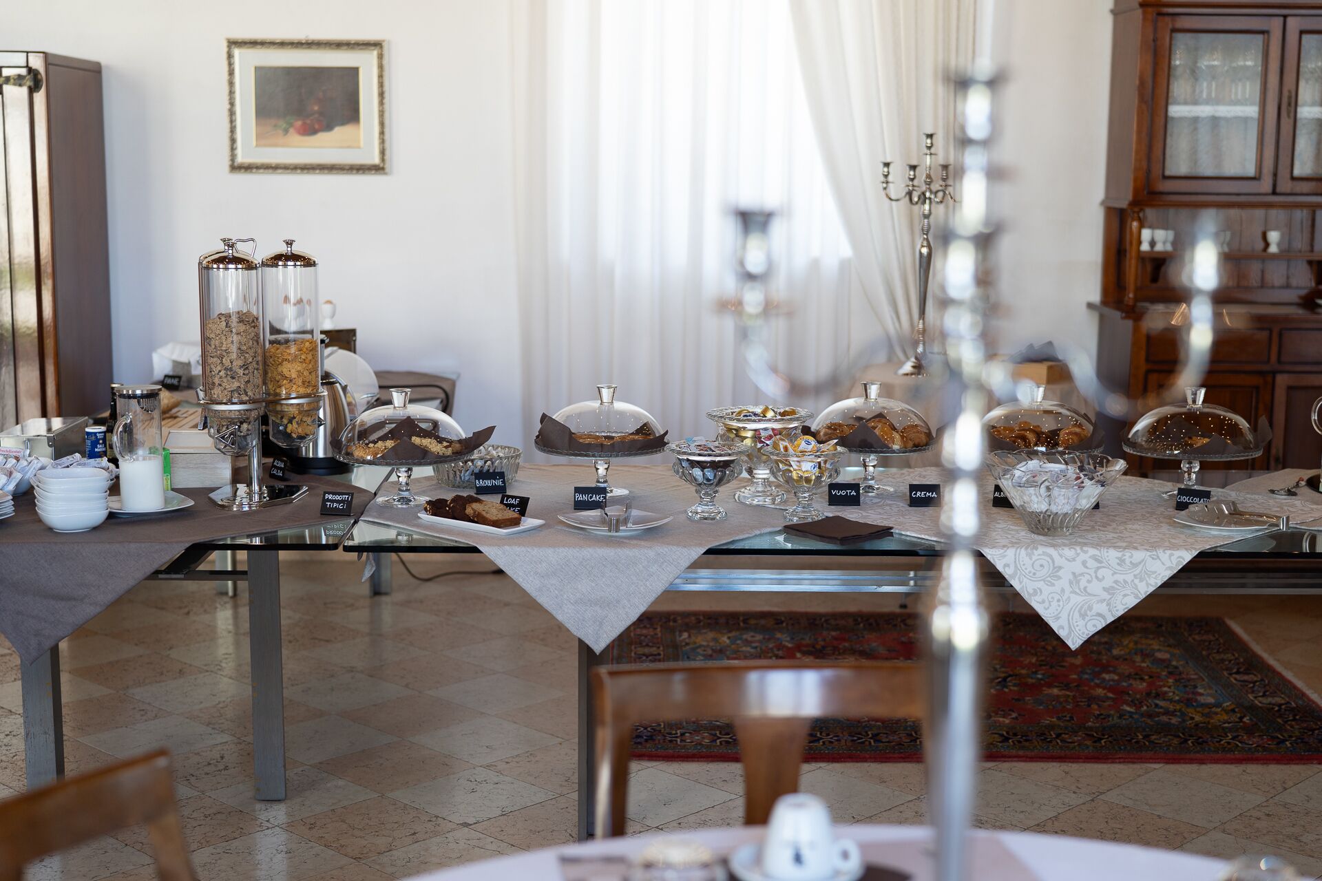 Zona pranzo con un'area buffet che presenta diversi alimenti su supporti in vetro, un lampadario e un tappeto decorato.