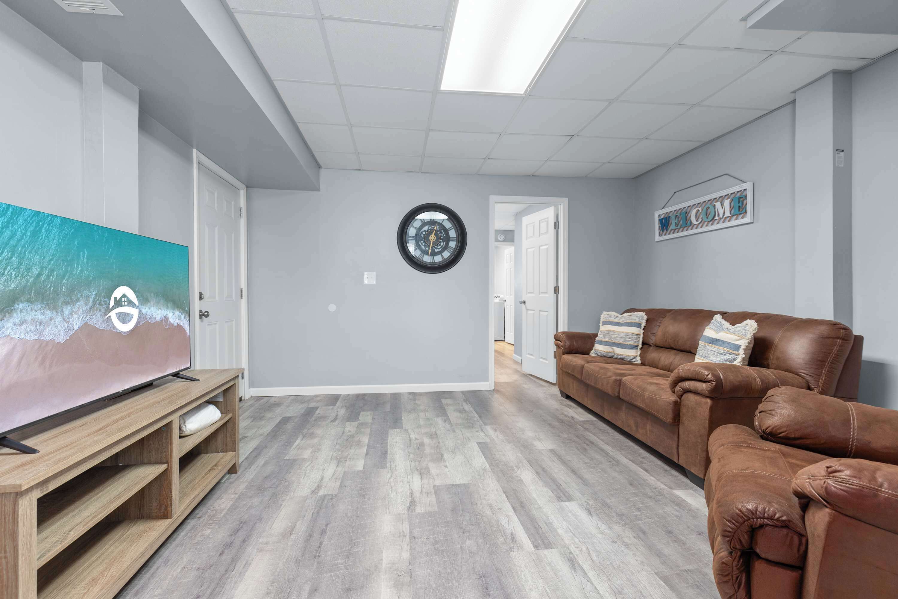 A living room with a flat-screen TV, a brown leather sofa, a wooden entertainment unit, and a 'WELCOME' sign on the wall.