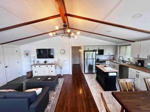 A spacious loft-style living area with a kitchen and dining area, featuring a high ceiling with exposed beams, a large TV, and a modern kitchen setup. - 8th Street Haven - House in DT Hamilton (Hamilton)